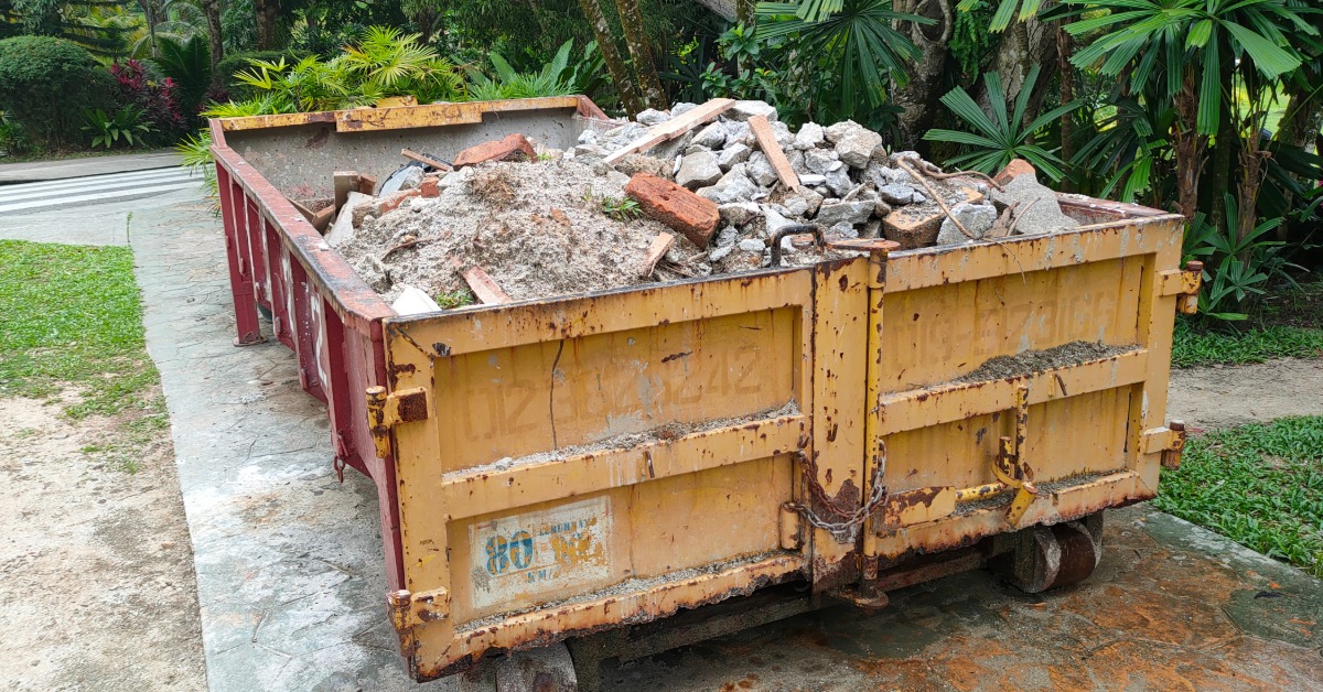 A medium size dumpster with yellow doors and red sides full of debris, placed on a driveway next to palm trees.